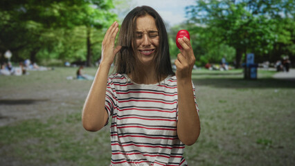 Fototapeta premium Woman holding small red ball with hand to head and squinting in forest park under trees and sunlight; pain headache.