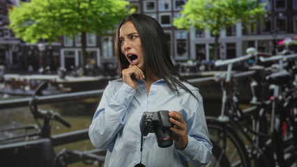 Fototapeta premium Woman holding camera and pulling at shirt while grimacing on street in amsterdam canal area; discomfort travel fatigue.