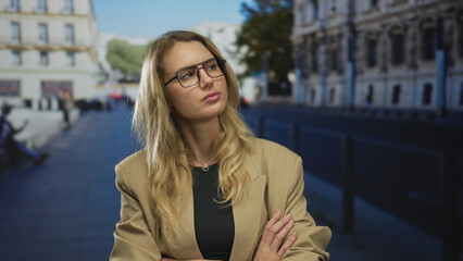 Fototapeta premium Woman standing on city street with arms crossed looking disgusted outdoor in an urban setting, featuring a young, blonde, caucasian woman in a blazer.