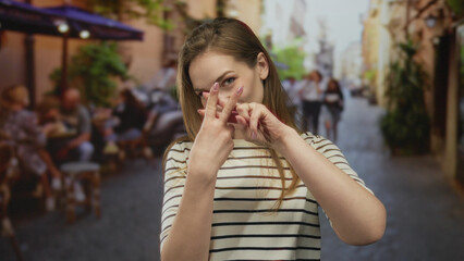 Fototapeta premium Woman crossing index fingers in front of her on a busy street terrace by cafe tables under umbrellas with a playful smile; rejection.