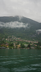 Obraz premium Lakeside village with waterfront buildings, rippling emerald lake water and low clouds clinging to the mountain slopes; serenity.