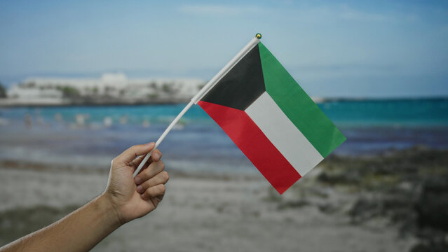 Man holding kuwaiti flag at seaside beach, showcasing national pride and cultural identity in outdoor setting under clear blue sky.