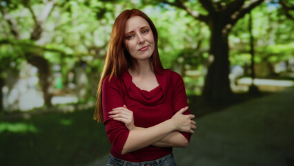 Obraz premium Woman standing in a sunny park with crossed arms, displaying an expression of curiosity and contemplation amidst lush green trees during a peaceful day.