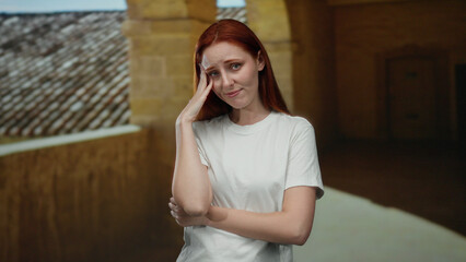 Fototapeta premium Woman standing in outdoor university courtyard with hand on head, appearing thoughtful and reflective, showcasing a serene and picturesque academic environment.