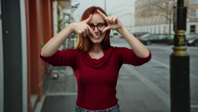 Woman posing on a city street, wearing glasses and a red sweater, using fingers to frame something, blonde hair, day scene, stylish fashion, thoughtful moment, urban backdrop.