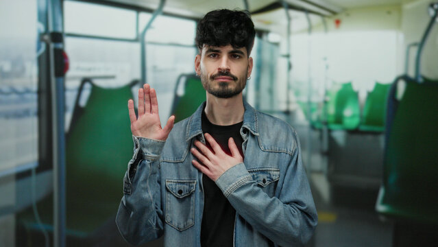 Young man making a pledge with hand on chest inside a bus, wearing a denim jacket, showcasing a thoughtful expression, demonstrating a solemn commitment in an urban transport setting.