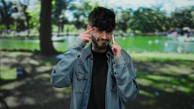 Young man talking on phone in a sunny park landscape wearing denim, expressing thoughtfulness with gestures.