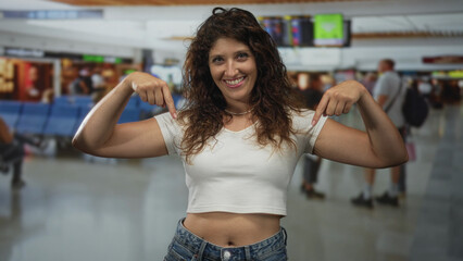 Young woman points both fingers to bare abdomen in busy airport terminal while smiling and wearing...