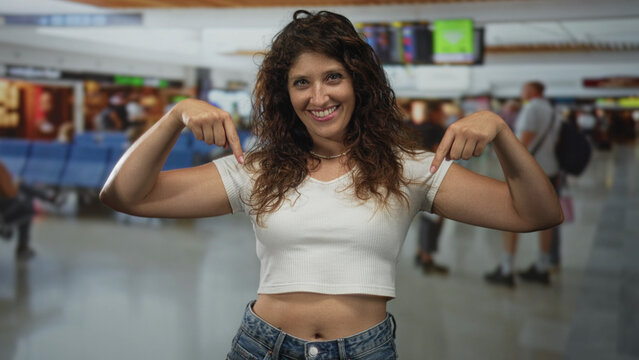 Young woman points both fingers to bare abdomen in busy airport terminal while smiling and wearing white crop top; travel confidence.