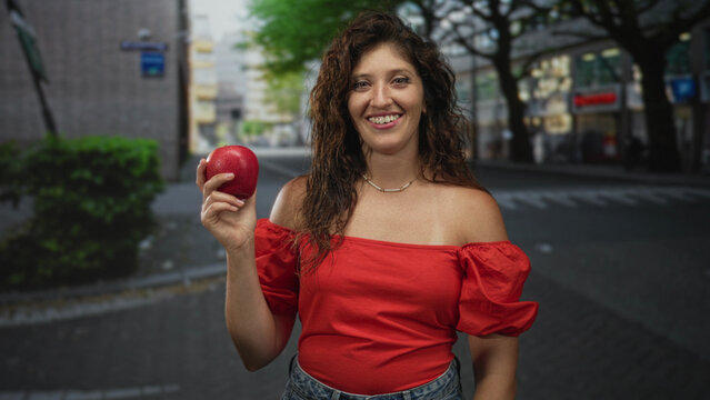 Woman holding red apple on a city street wearing denim and a necklace, smiling with exposed shoulders and hands visible; happiness.