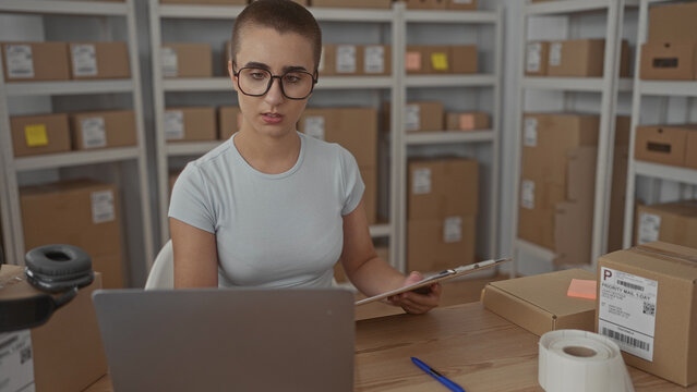 Woman holding clipboard and writing with pen with hand on table amid shipping boxes in building; focused entrepreneurship.