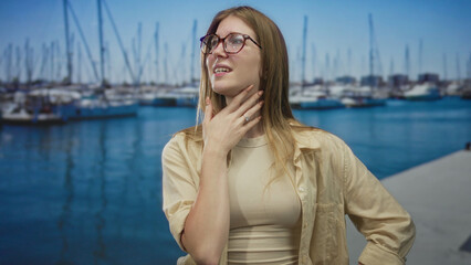 Fototapeta premium Blonde woman wearing glasses pinches nose beside boats on sunlit street pier over blue water; disgust.