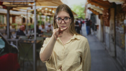 Obraz premium Blonde woman holds hand making v sign at outdoor restaurant terrace and looks at camera with serious expression; defiance.