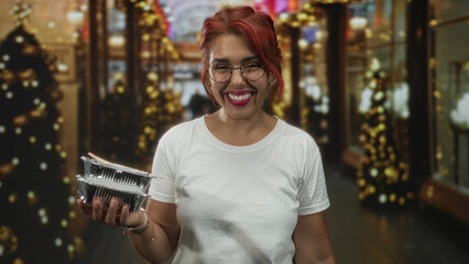 Fototapeta premium Woman holding aluminum food containers and smiling while wearing glasses and a white tshirt on decorated street with holiday lights; community sharing joy.