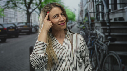 Fototapeta premium Blonde middle aged woman with long hair wearing striped shirt pointing finger to head on green street; thinking.