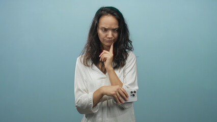 Obraz premium Woman holding smartphone with finger to chin and examining phone screen in studio; uncertainty hesitation doubt.
