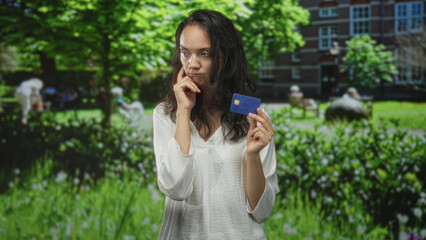 Fototapeta premium Woman holding blue creditcard with palm up questioning in building courtyard; uncertainty decision hesitation.
