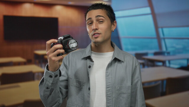 Young man holding camera in classroom, showcasing expressions and gestures in an indoor setting, emphasizing education, photography, learning, and creativity.