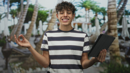 Fototapeta premium Teenage boy holding tablet and making ok sign beside palm trees and pool at resort building; relaxation mindfulness.