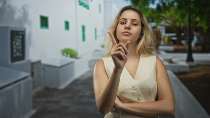 Woman points index finger upward on street in front of white building, arms crossed with bare shoulder visible, direct gaze at camera; confidence warning.