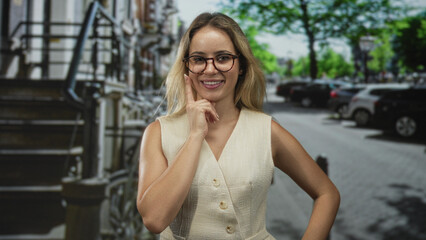 Fototapeta premium Woman with hand on cheek on street wearing glasses and sleeveless beige vest, smiling in urban outdoors; thoughtful curiosity.