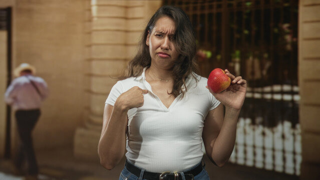 Woman holding red apple pointing finger to chest by building entrance; skepticism healthy eating choice.