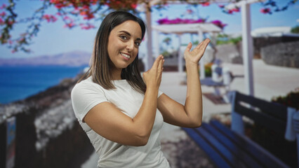 Fototapeta premium Young hispanic woman in white tshirt points fingers upward and shows palms while smiling on a studio seaside mural set; welcoming joy.