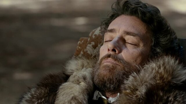 A man lies back on the ground wrapped in warm clothing. He enjoys the sun while surrounded by snow-covered ground and trees. This moment captures the winter leisure.