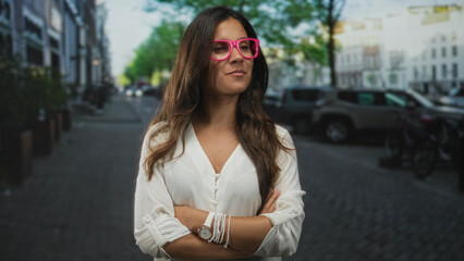 Obraz premium Woman wearing pink glasses with arms crossed on a city street near parked cars and bicycles; confidence determination poise independence.