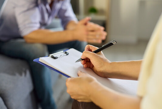 Psychologist and patient talk in therapy session. Clinician writes notes on a clipboard as a man talks about mental health during consultation. Concept: mental health counseling process.
