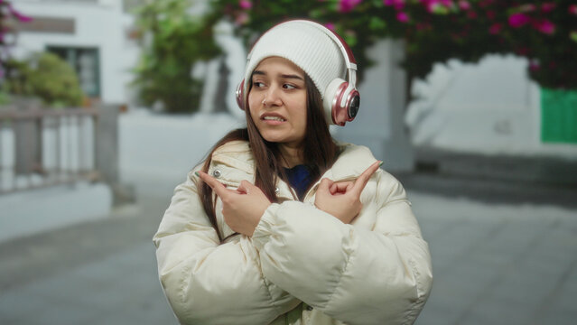 Young woman wearing headphones and a winter jacket listens to music on a peaceful street lined with flowering trees, enjoying a moment outdoors.