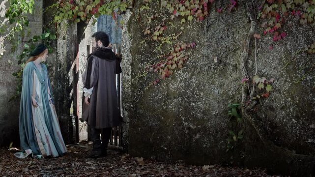A couple walks through a stone wall covered in vines. They stop to look at a wooden gate. The leaves on the ground show that it is autumn. The setting feels old and mysterious.