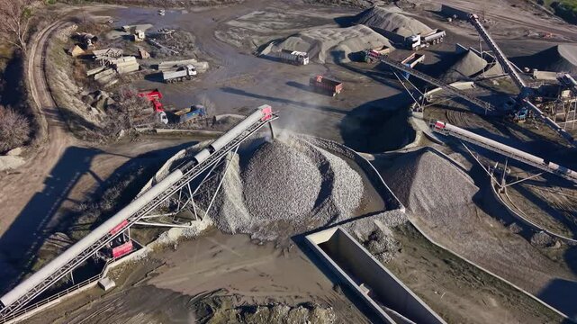 Heavy machinery works tirelessly at a stone quarry, shaping the landscape with towering piles of gravel and pathways stretching under the clear sky. Dust swirls as trucks transport materials.