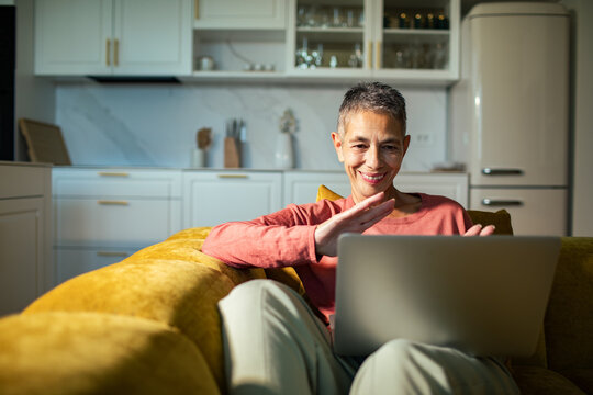 Senior woman video chatting on laptop on sofa in home kitchen