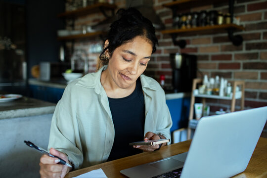 Woman budgeting with smartphone and laptop at home kitchen