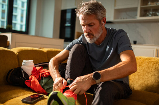 Middle-aged man tying running shoes on living room sofa