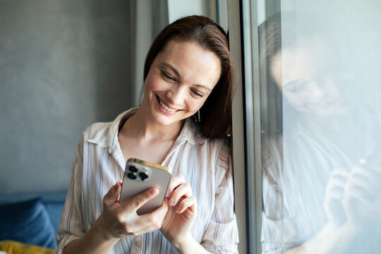 Smiling woman texting on smartphone by window at home