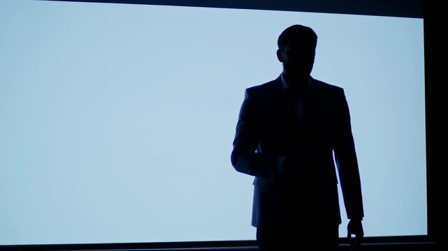 A man in a business suit standing alone on a stage with a screen behind him