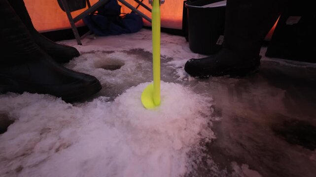 Low perspective inside tent shows two black boots, three drilled ice holes, warm orange tint on ice, slight rod jig over center hole, faint water ripples, winter scene.