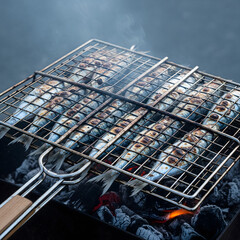Fototapeta premium Row of fresh sardines with charred skin grilling over hot charcoal in a metal basket with smoke rising against a dark background. Generative AI