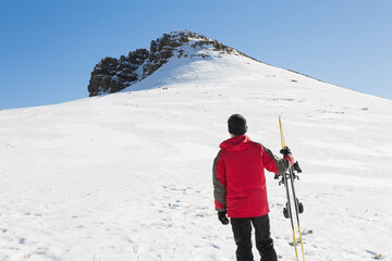 Skier is standing on broad snowfield holding skis and poles near rocky peak, clear sky