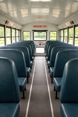 Obraz premium Empty school bus interior with blue vinyl seats and central aisle looking toward rear emergency exit safety sign bright daylight through windows public transportation and student commute