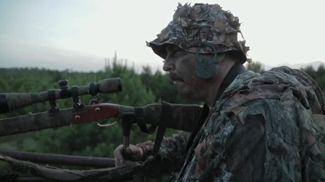 A hunter waits quietly in the forest, dressed in camouflage, as he aims his weapon. The sun sets behind the mountains, creating shadows in the trees.