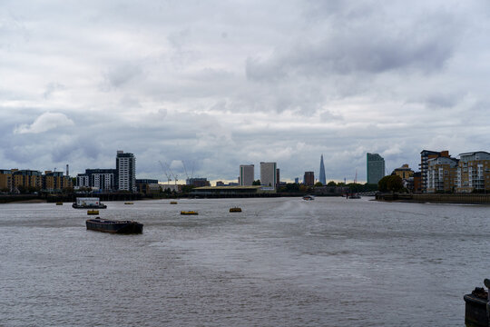 Cloudy London cityscape along the Thames, featuring the city skyline, The Shard, and boats on the river.