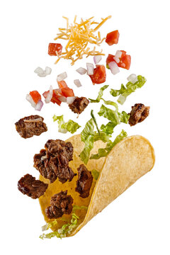 deconstructed beef taco with flying ingredients: seasoned meat, shredded lettuce, diced tomatoes, onions, and cheese over a corn tortilla, isolated.