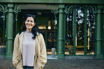 Naklejka premium Smiling woman stands outdoors in front of a green ornate building, wearing a beige jacket and gray sweater, candid urban scene with natural light.