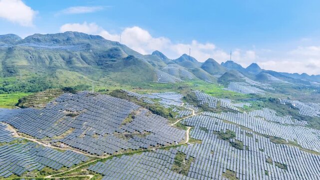 Aerial time-lapse of a large solar power station on rocky desertified mountains with rows of photovoltaic panels covering the terrain and wind turbines on mountain ridges, new energy landscape.
