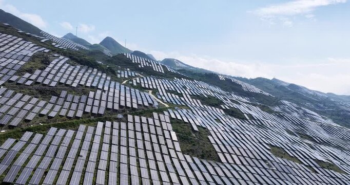 Aerial footage of solar photovoltaic farm on rocky hills, rising perspective showcasing renewable energy in mountainous terrain under clear sky. 