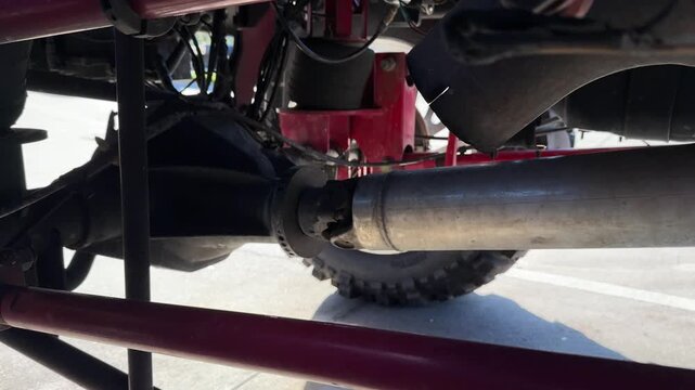 Detailed view shows a truck axle and driveshaft under the vehicle chassis. Heavy mechanical components, rods and joints highlight the rugged undercarriage of the automotive suspension system.