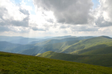 Vast green hills roll into distant mountains beneath a dramatic, cloud filled sky. Soft sunlight breaks through, creating peaceful, natural scenery. Carpathian Mountains, Ukraine 